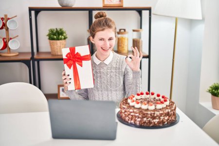 Photo for Cheerful young blonde woman joyously celebrating her birthday over a video call. gesturing an 'okay' sign, with a friendly smile, showcasing her gift while comfortably at home. - Royalty Free Image