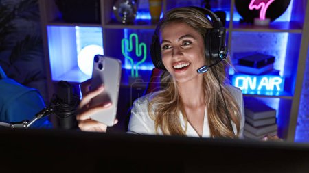 Foto de Atractiva joven mujer streamer rubia captura selfie sala de juegos de noche, sonriendo a la tecnología de teléfonos inteligentes portátiles en medio del entretenimiento digital - Imagen libre de derechos
