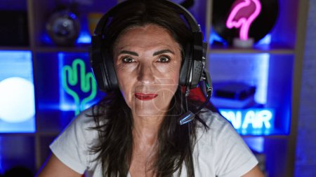 Photo for A mature woman with headphones in a gaming room lit by colorful neon lights at night, portraying a vibrant yet focused indoor scene. - Royalty Free Image