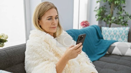 Photo for A mature woman wrapped in a cozy blanket uses a smartphone in a modern living room interior. - Royalty Free Image