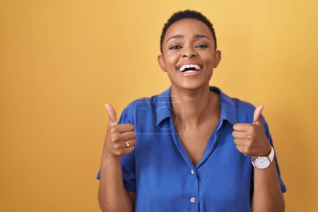 Téléchargez les photos : Femme afro-américaine debout sur fond jaune signe de succès faisant geste positif avec la main, pouces levés souriant et heureux. expression joyeuse et geste gagnant. - en image libre de droit