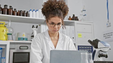Foto de Mujer hispana con cabello rizado usando gafas y bata de laboratorio trabaja en interiores en una computadora de laboratorio, rodeada de equipo. - Imagen libre de derechos