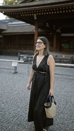 Téléchargez les photos : Posant en toute confiance à fushimi inari taisha, une belle femme hispanique portant des lunettes, sourit largement, regardant autour de lui, captivée par les magnifiques environs du temple kyoto. - en image libre de droit