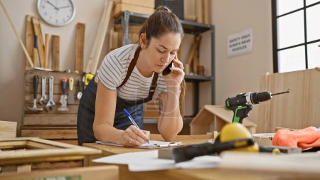 Photo for A focused woman in a carpentry workshop talks on the phone while taking notes amidst tools and woodwork. - Royalty Free Image