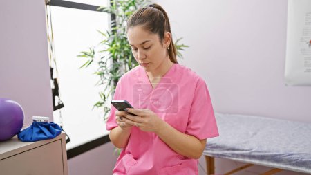 Téléchargez les photos : Une jeune femme en blouse rose utilisant un smartphone dans une salle de clinique de physiothérapie lumineuse. - en image libre de droit