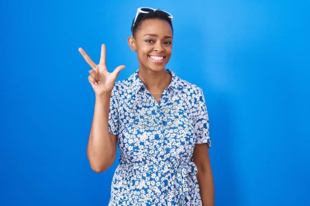 Téléchargez les photos : Femme afro-américaine debout sur fond bleu montrant et pointant vers le haut avec les doigts numéro trois tout en souriant confiant et heureux. - en image libre de droit