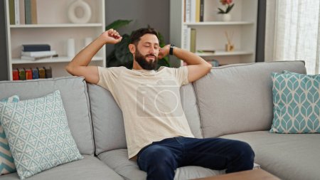 Téléchargez les photos : Jeune homme hispanique assis sur le canapé fatigué bras tendus à la maison - en image libre de droit