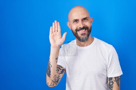 Foto de Hombre hispano con tatuajes de pie sobre fondo azul renunciando a decir hola feliz y sonriente, gesto de bienvenida amistoso - Imagen libre de derechos