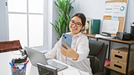 Foto de Una alegre joven hispana se toma una selfie en su escritorio de oficina, rodeada de tecnología y artículos de trabajo. - Imagen libre de derechos