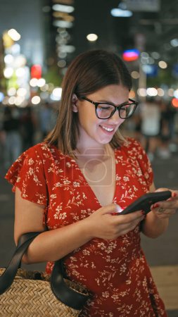 Photo for Cheerful and beautiful hispanic woman, captured in a joyful moment, texting on her smartphone under the night lights of urban osaka street, embodying the essence of a connected digital world. - Royalty Free Image