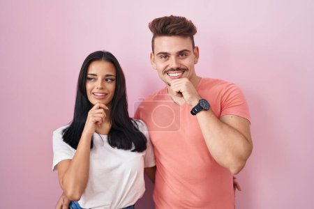 Téléchargez les photos : Jeune couple hispanique debout sur fond rose avec la main sur le menton penser à la question, expression coûteuse. Visage souriant et réfléchi. concept de doute. - en image libre de droit