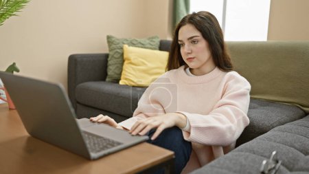 Foto de Una joven contemplativa que usa una computadora portátil en una acogedora sala de estar captura el estilo de vida moderno del trabajo desde el hogar. - Imagen libre de derechos