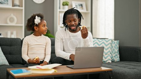 Téléchargez les photos : Père et fille afro-américains assis sur le canapé manger croissant dessin et travailler à la maison - en image libre de droit