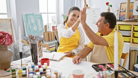 Photo for Hispanic man and woman artists high five in joy at art studio while drawing on notebook; vibrant celebration amidst paintbrushes, easels, and canvas around - Royalty Free Image