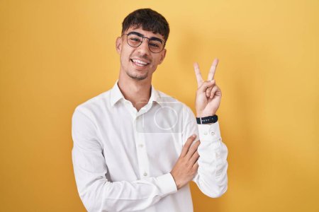 Photo for Young hispanic man standing over yellow background smiling with happy face winking at the camera doing victory sign with fingers. number two. - Royalty Free Image
