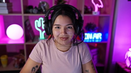 Photo for Smiling young hispanic woman with headphones in a colorful gaming room at night. - Royalty Free Image