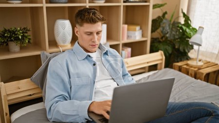 Photo for Handsome young caucasian man, relaxed in curated indoor morning light, sitting comfortably on cozy bed, fully awake, his serious expression shows concentration as he uses laptop at home - Royalty Free Image