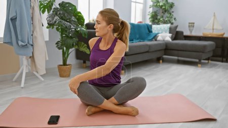 Photo for A middle-aged woman practices yoga in a well-lit living room, next to a smartphone on a pink mat. - Royalty Free Image