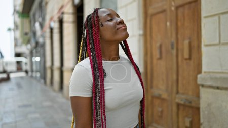 Téléchargez les photos : Belle femme afro-américaine profitant de l'air frais, les yeux fermés, accueillant souffle d'été de la rue de la ville tout en se tenant debout, méditant de façon décontractée au milieu de l'extérieur urbain sous le soleil ensoleillé - en image libre de droit