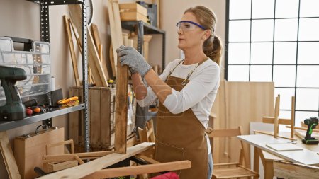 Téléchargez les photos : Femme d'âge moyen artisan inspecte le bois dans un atelier ensoleillé - en image libre de droit