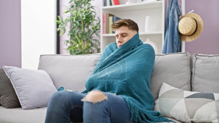 Young caucasian man sitting on sofa covering blanket for cold at home
