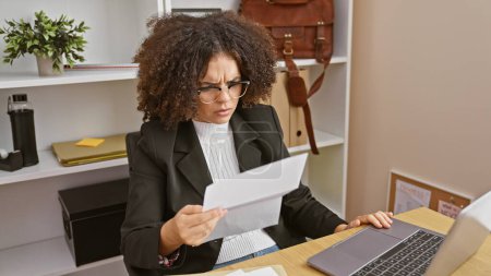 Foto de Una joven hispana con el pelo rizado, gafas y una chaqueta lee un documento en una oficina moderna. - Imagen libre de derechos