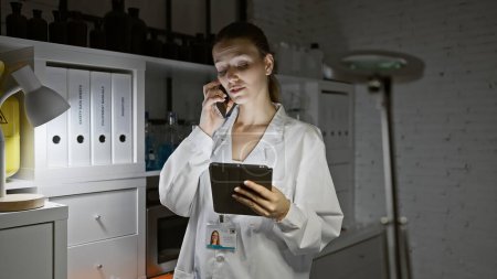 Photo for A young woman in a lab coat multitasking in a pharmacy with a phone and tablet. - Royalty Free Image