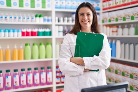 Foto de Joven bella mujer hispana farmacéutica sonriendo confiada sosteniendo portapapeles en la farmacia - Imagen libre de derechos