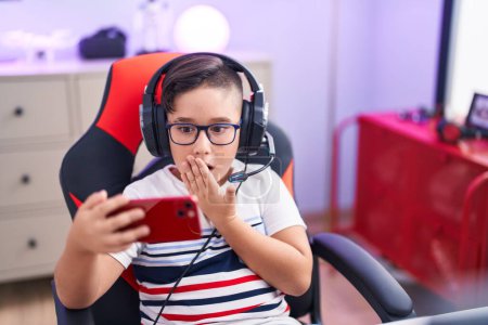 Téléchargez les photos : Jeune enfant hispanique jouant à des jeux vidéo avec smartphone couvrant la bouche avec la main, choqué et effrayé par l'erreur. expression surprise - en image libre de droit