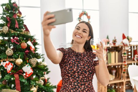 Téléchargez les photos : Young beautiful hispanic woman make selfie by smartphone standing by christmas tree at home - en image libre de droit