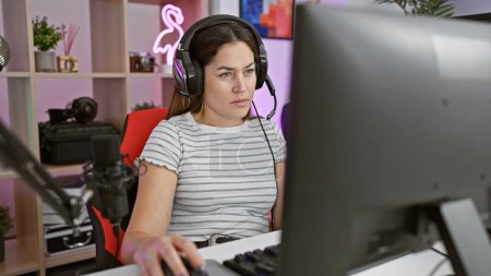 Foto de Mujer joven enfocada con auriculares usando una computadora en una moderna sala de juegos por la noche. - Imagen libre de derechos