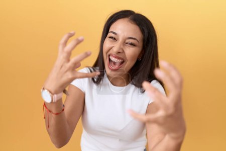 Photo for Young arab woman wearing casual white t shirt over yellow background shouting frustrated with rage, hands trying to strangle, yelling mad - Royalty Free Image