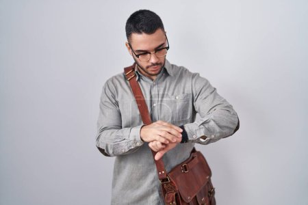 Foto de Joven hombre hispano con maleta revisando la hora en el reloj de pulsera, relajado y confiado - Imagen libre de derechos