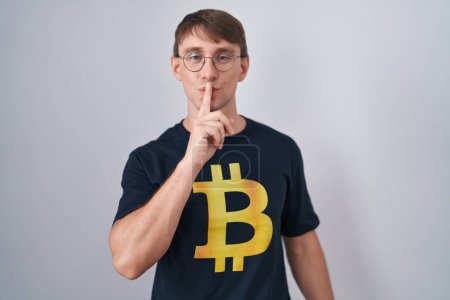 Photo for Caucasian blond man wearing bitcoin t shirt asking to be quiet with finger on lips. silence and secret concept. - Royalty Free Image