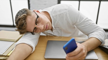 Foto de Agotado joven hombre de negocios caucásico, teléfono inteligente en la mano, pierde la batalla con el estrés noblemente se estrella dormido en la mesa de la oficina - Imagen libre de derechos