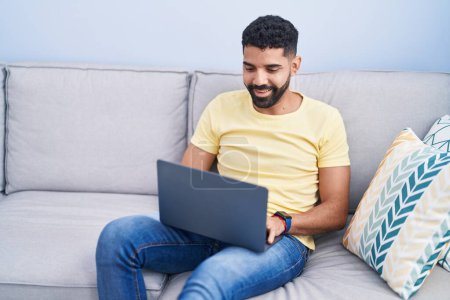 Téléchargez les photos : Jeune homme arabe utilisant un ordinateur portable assis sur la table à la maison - en image libre de droit