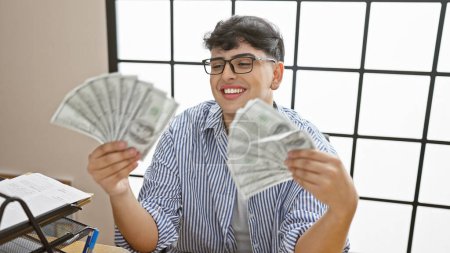 Foto de Joven sonriente con gafas sosteniendo dólares en la oficina moderna, irradiando éxito y felicidad. - Imagen libre de derechos