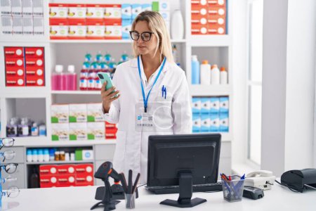 Photo for Young blonde woman pharmacist using smartphone and computer at pharmacy - Royalty Free Image