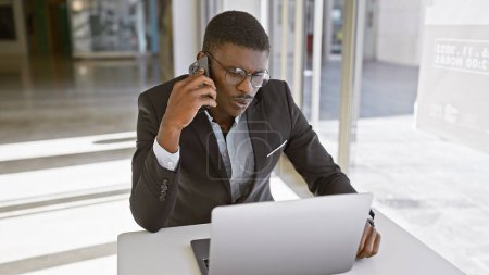 Téléchargez les photos : Bel homme africain en costume travaillant sur ordinateur portable et parlant au téléphone dans l'intérieur de bureau moderne. - en image libre de droit