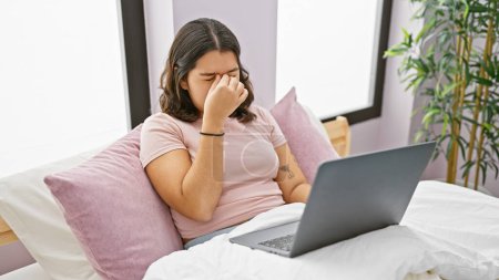 Téléchargez les photos : Une jeune femme hispanique semble stressée en utilisant un ordinateur portable dans sa chambre minimaliste. - en image libre de droit