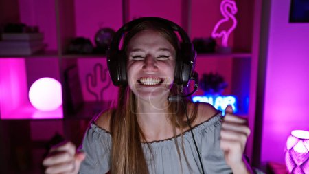 Photo for Young blonde woman with headphones laughing joyfully in a neon-lit gaming room at night. - Royalty Free Image