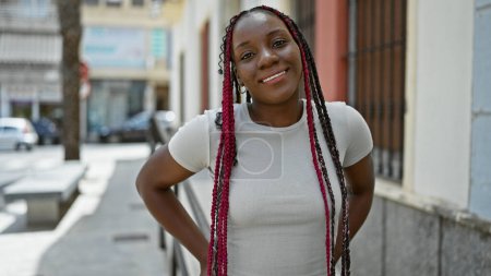 Téléchargez les photos : Belle femme afro-américaine riant, apportant de la joie dans la rue de la ville avec son sourire confiant et son style décontracté, rayonnant d'énergie positive sous le ciel ensoleillé - en image libre de droit