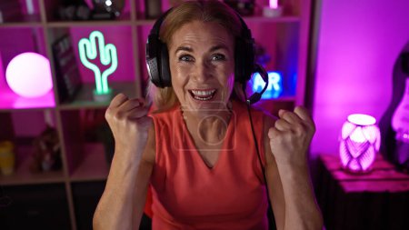 Photo for Excited mature woman with headphones in neon-lit gaming room celebrating victory at night. - Royalty Free Image