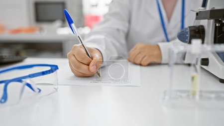 Photo for Professional woman in lab coat writing on a form, with microscope and eyeglasses in a clinical laboratory setting. - Royalty Free Image