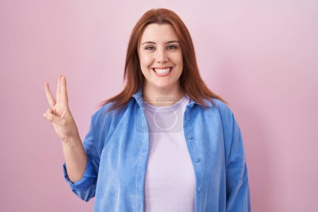 Téléchargez les photos : Jeune femme hispanique aux cheveux roux debout sur fond rose montrant et pointant vers le haut avec les doigts numéro trois tout en souriant confiant et heureux. - en image libre de droit