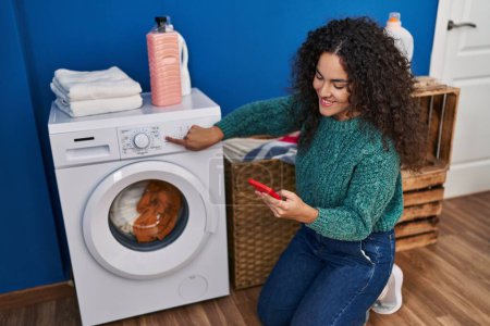 Photo for Young beautiful hispanic woman using smartphone turning on washing machine at laundry room - Royalty Free Image