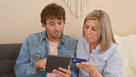 Relaxed mother and son bonding together at home, casually sitting on the sofa, speaking about tech-ridden lifestyle, using a touchpad for buying stuff, paying with a credit card.