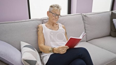 Photo for Cozy at home, senior grey-haired woman in glasses relaxing with a book on the sofa - Royalty Free Image