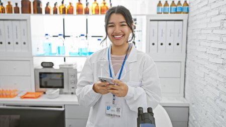 Photo for A smiling young hispanic woman in a lab coat uses a smartphone in a clinical laboratory setting. - Royalty Free Image