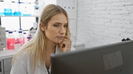 Téléchargez les photos : Sérieuse, belle jeune femme blonde scientifique réfléchissant fort tout en travaillant sur ordinateur, effectuant des recherches dans un laboratoire médical - en image libre de droit
